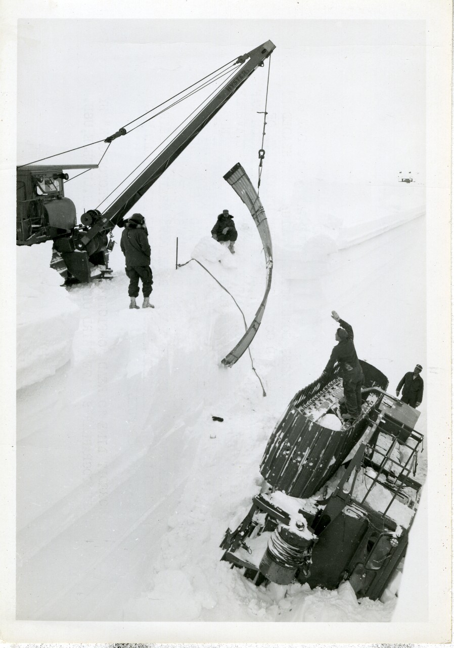 Black and white photograph of members of Task Force 43 use a cable hoist on a D-8 tractor to lift the 14-foot arches off of the tractor that fell into the trench, New Byrd Station, Antarctica, 21 January, 1961