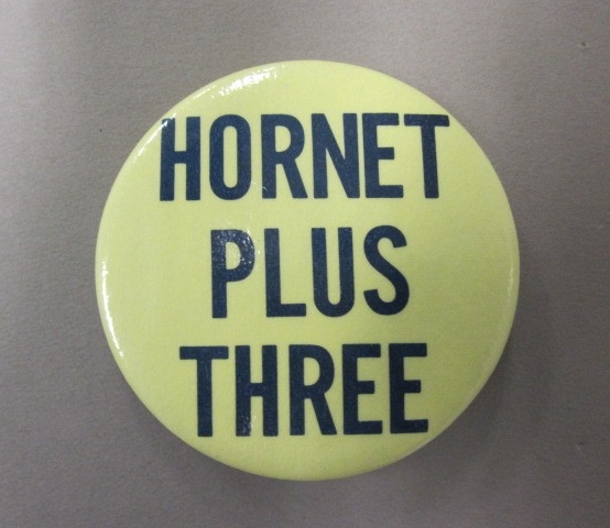 One circular yellow metal button with a pin clasp on the reverse side and dark blue lettering on the obverse side that reads  "Hornet / Plus / Three."