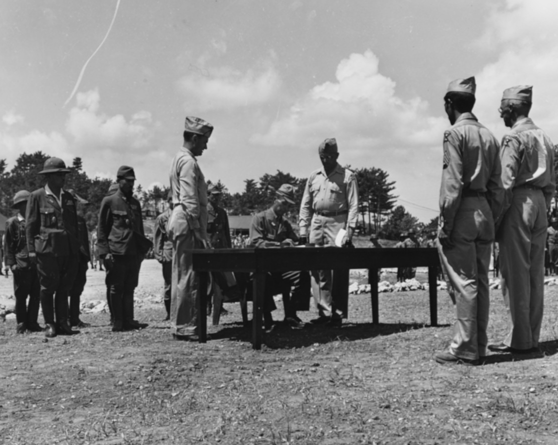 A Japanese offcer signs the surrender document on Okinawa, 7 September 1945. Also at table, standing (left to right) are Col. Phillip Bethune and Maj. Gen. Frank Merrill. At right is also General Joseph Stillwell (80-G-344922).