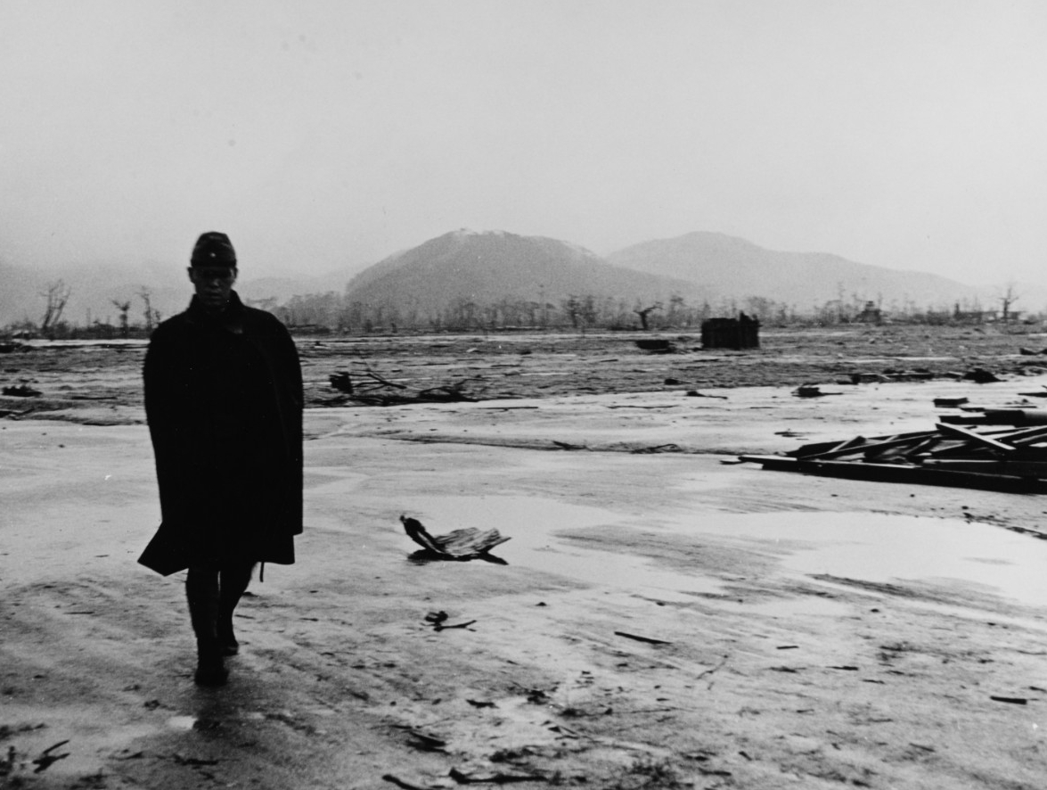 Black-and-white photograph of a Japanese soldier walking toward the viewer, silhouetted against a background that shows the flattened city of Hiroshima and scattered debris. In the distant background are burnt trees and mountains.