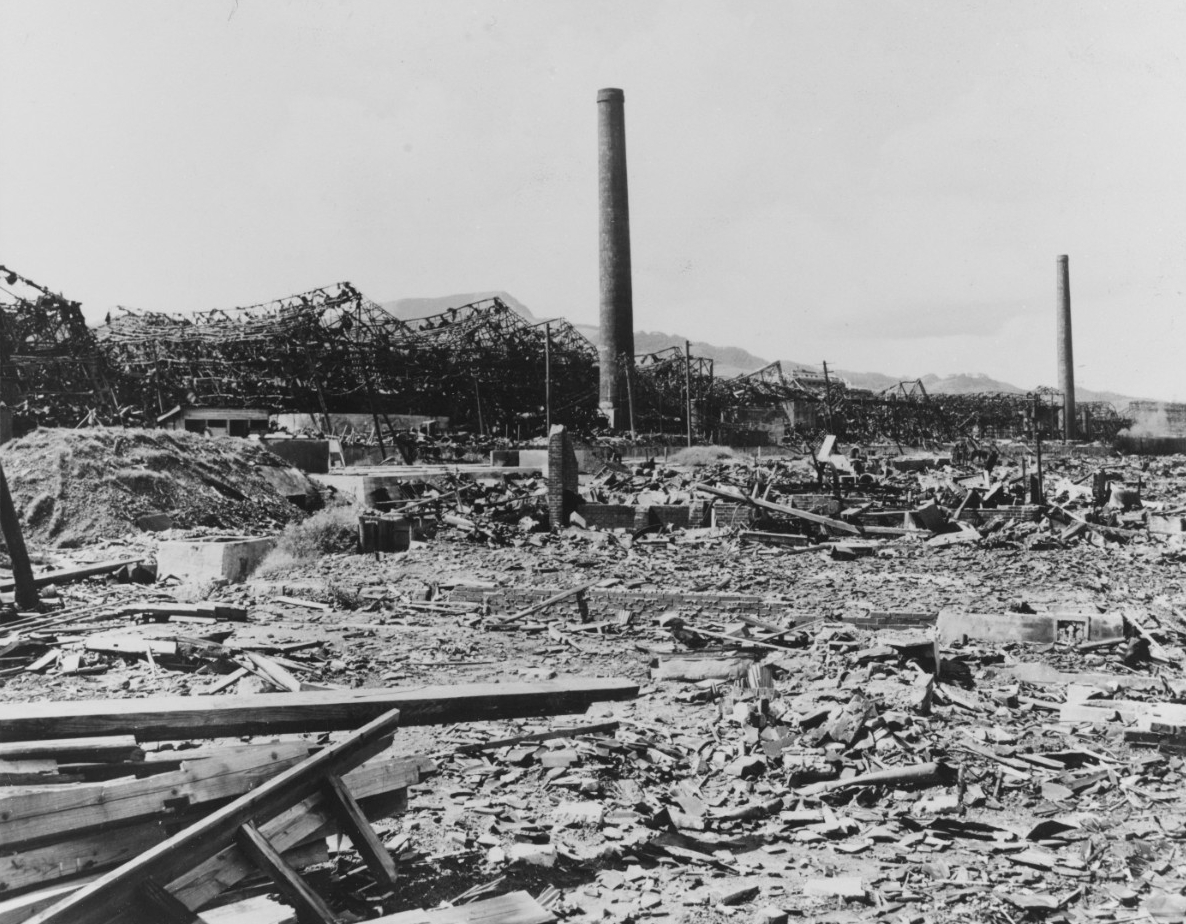 A ground-level view of damage to what appears to have been an industrial site in Nagasaki, Japan. The ground is littered with debris, with planks of wood in the lower left. In the background stands a series of ruins and two smokestacks, still sta...