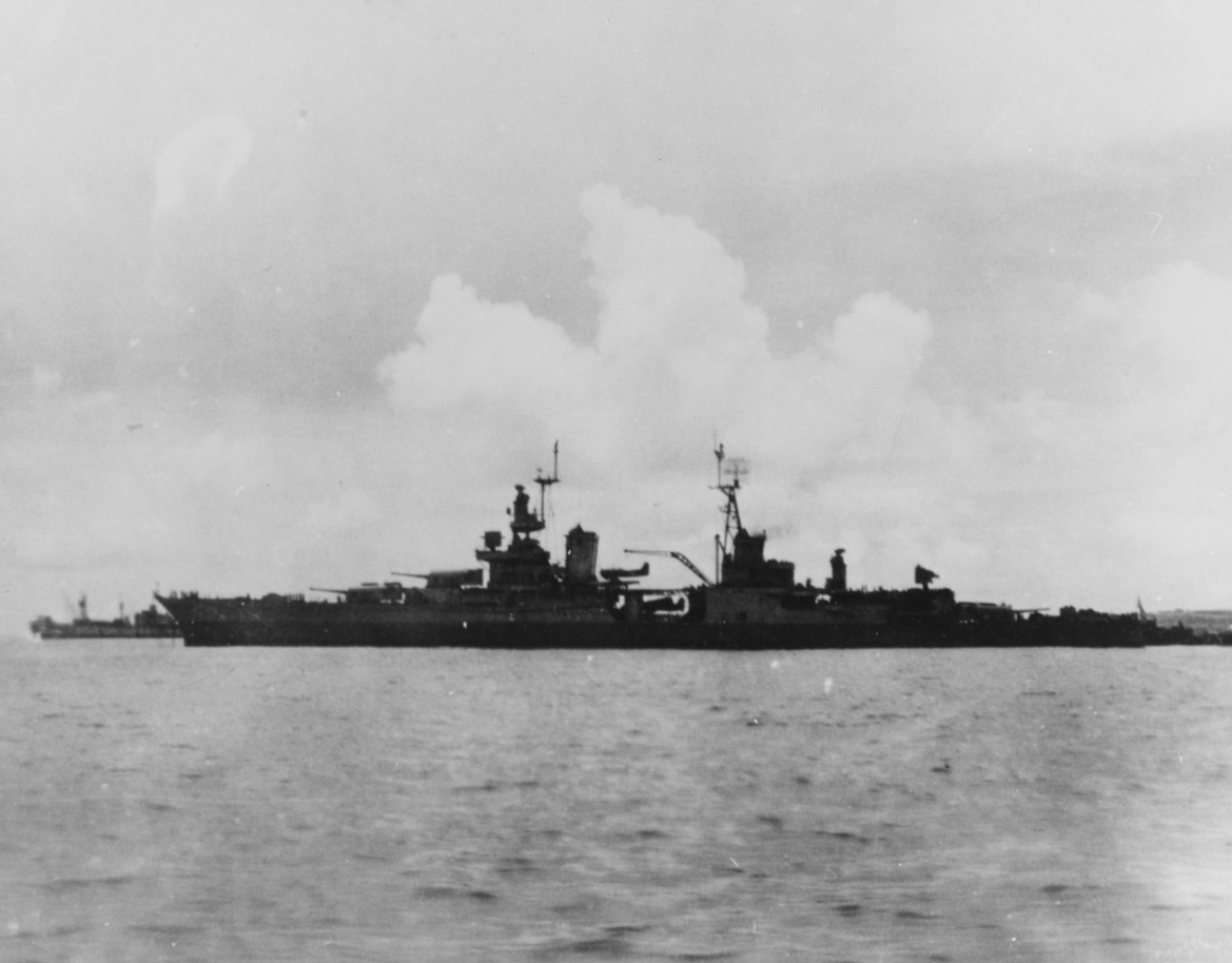Black-and-white photograph seen from the side, backlit, with clouds, another ship, and land in the background.