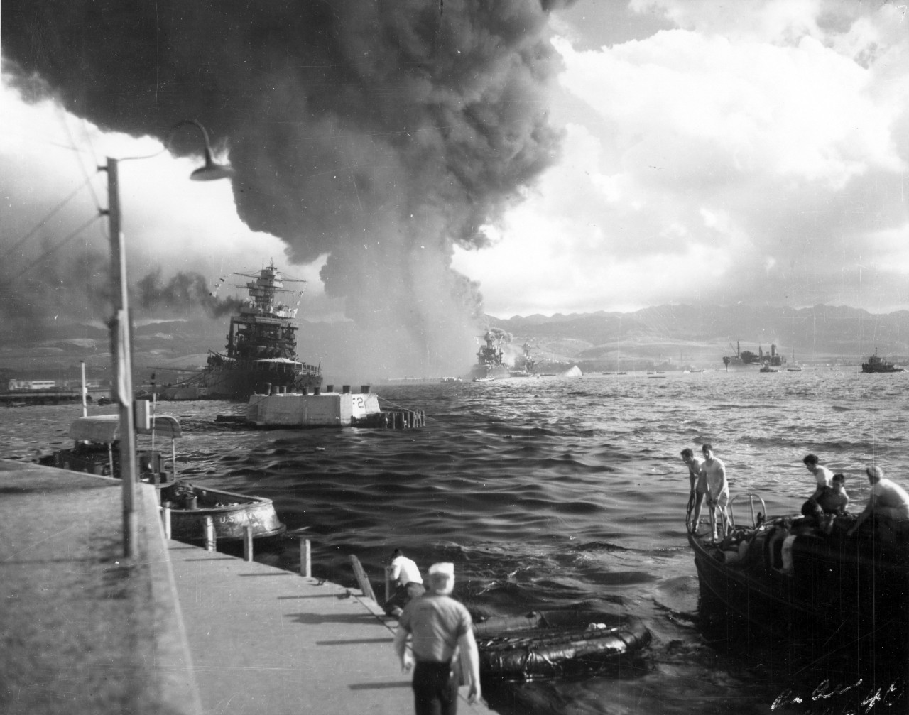 View of harbor with stricken, sinking ships and visible smoke.