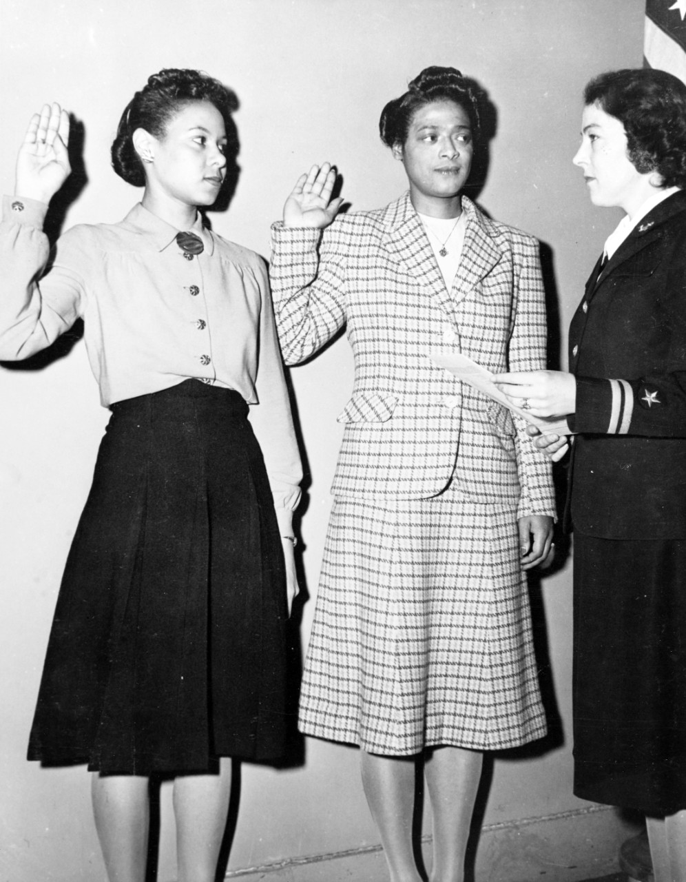 Two women hold up their right hand while looking at a female Navy lieutenant.