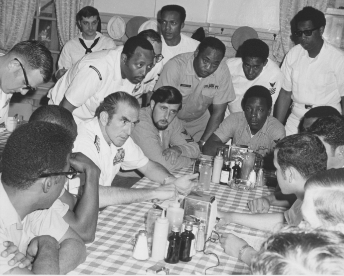 Admiral Zumwalt meeting with Sailors. Admiral Zumwalt and Sailors are seated at a table with other Sailors standing around the table.