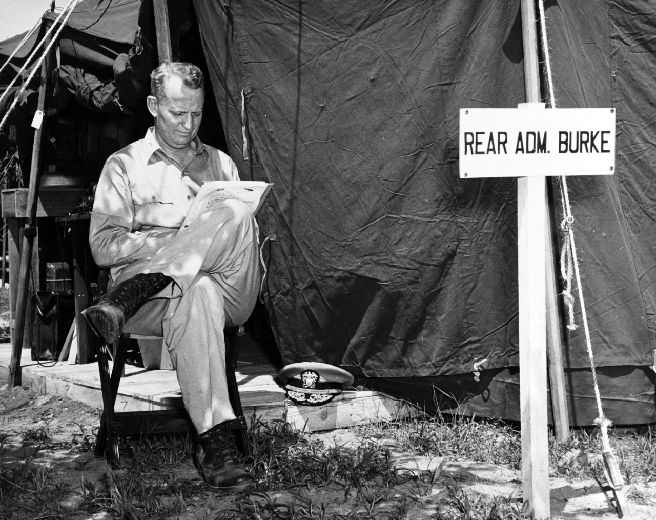 photo of man seated in front of a tent that bears the sign "Rear Adm. Burke" reading papers with a serious expression