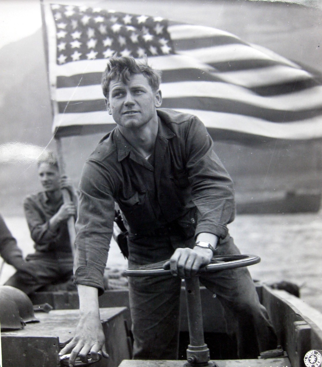 With the American flag flying over his head, Motor Machinist’s Mate Robert Mooty, USN, ferries troops across the Rhine River at Oberwesel, Germany.