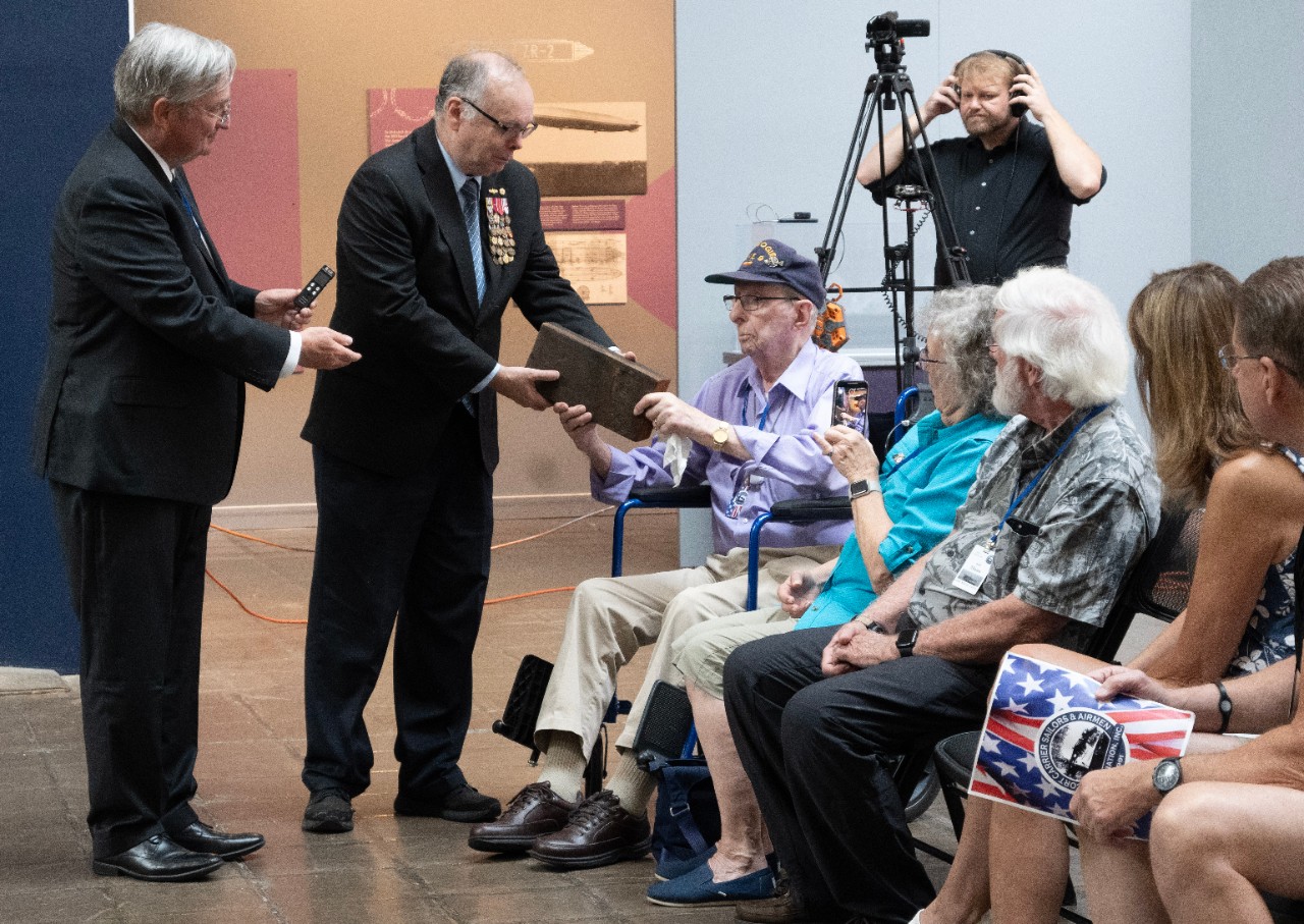 Robert Kruger, 100-year-old WWII veteran offers Naval History and Heritage Command Director Samuel Cox a plank of wood from the flight deck of USS Bogue (CVE 9) where he served in WWII. The Escort Carriers Sailors and Airmen Association (ECSAA) v...