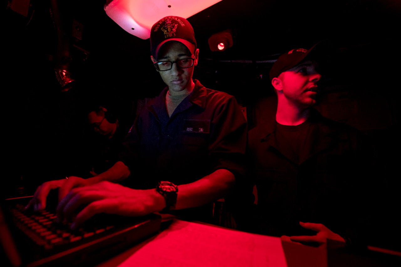 Sailor wearing USS Kearsarge cap entering information into a computer while standing next to another Sailor, in a room illuminated by a red light.