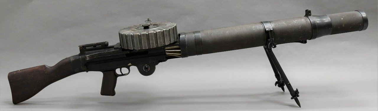 Right side of an American Lewis machine gun. The barrel is a large diameter tube with fins at the rear. Brown wood pistol grip and buttstock. The barrel is propped up by a metal bipod behind the muzzle. A circular pan magazine sits atop the receiver.