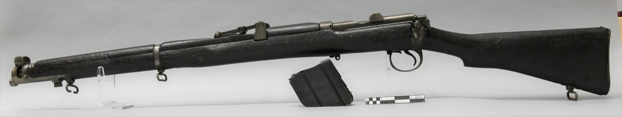 Left side of Lee-Enfield No. 1 Mk 3* Short Magazine rifle. Rifle has full-length dark wood stock with hooked semi-pistol grip. Metal triangular magazine unloaded and sitting in next to rifle.