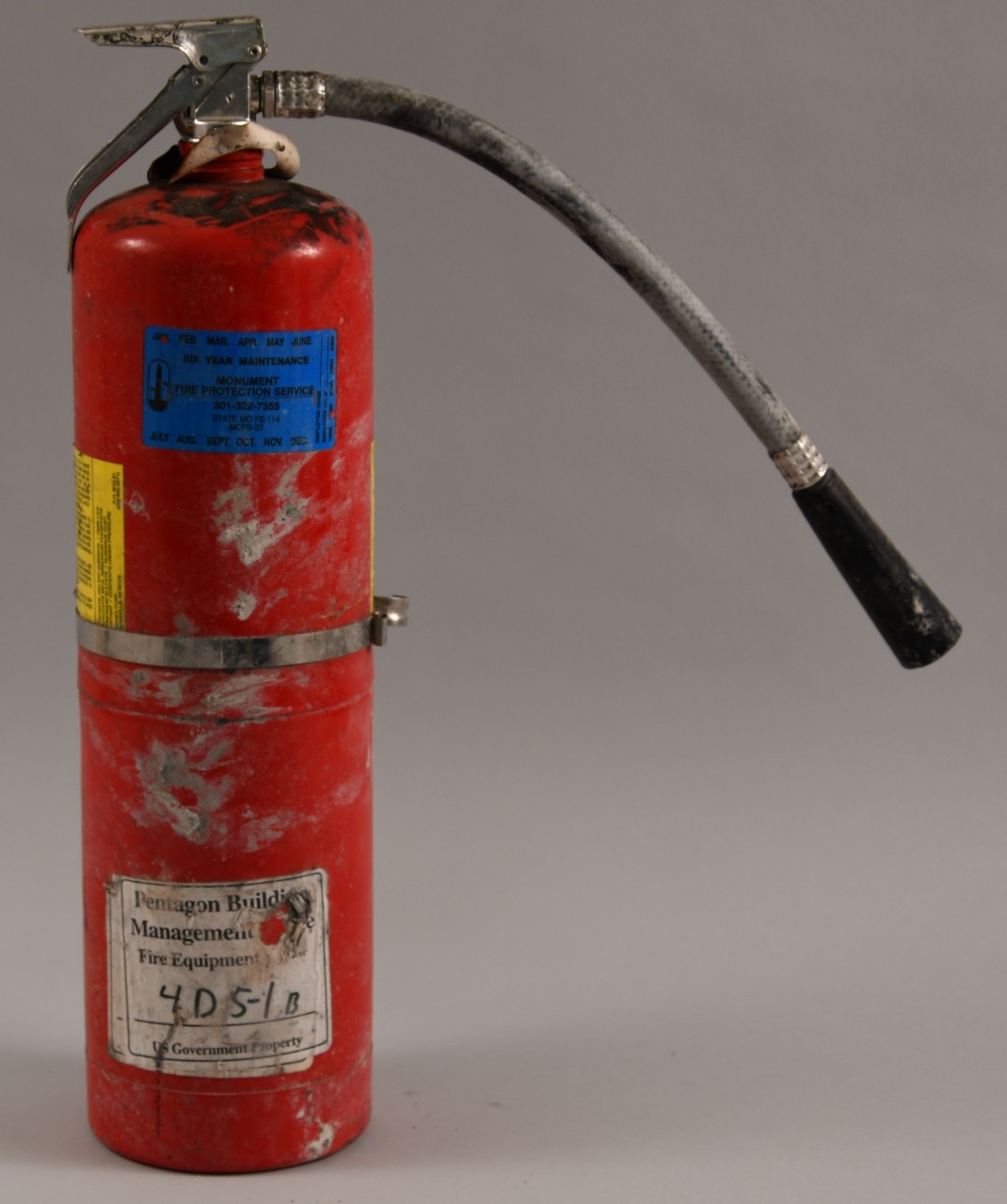 Reverse side of red fire extinguisher, hose extended to the right. White property label affixed to lower section. 