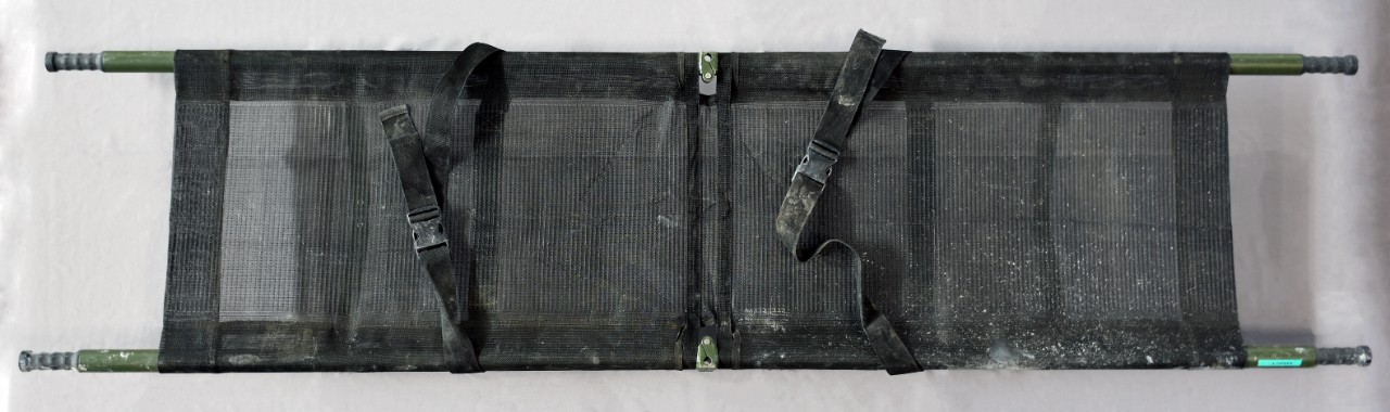 Obverse side of foldable black mesh stretcher. Two securing straps across body. Two carrying poles.