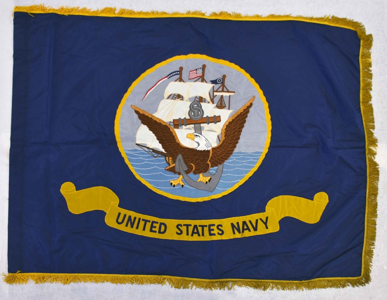 One US Navy flag. The flag is rectangular-shaped blue nylon edged with gold fringe. At the center of the flag is the seal of the US Navy with the eagle, anchor, and sailing ship above a banner with the words “United States Navy.” There are smoke ...
