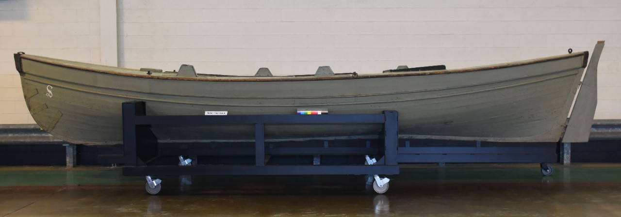 Full port view of small wood boat on mobile metal carriage. Boat painted gray with white painted S on bow. Wood tiller on stern.