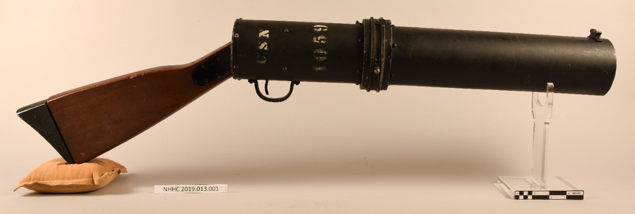 Obverse view of blinker tube/signal light gun. Black metal tube with white text, trigger on underside, wood gun stock.