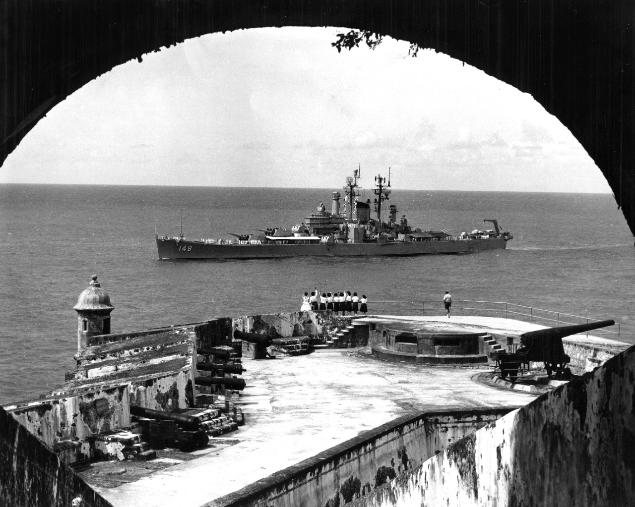 1 black and white photograph of USS Newport News (CA-148) passing Fort San Cristobal, Puerto Rico, donated by John Williams.