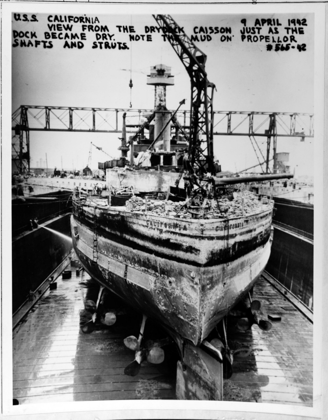 California enters Dry Dock No. 2 at Pearl Harbor for repairs, 9 April 1942. Shipyard workers have just drained the water from the dry dock, exposing the mud on her propeller shafts and struts. (Unattributed U.S. Navy Photograph NH 64483, Photographic Section, Naval History and Heritage Command)