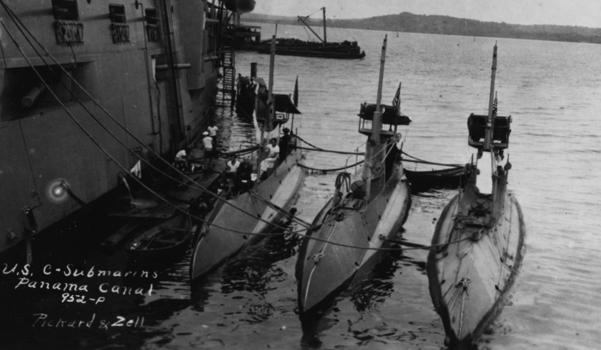 Charleston tends (left–right) C-5 (Submarine No. 16), C-2 (Submarine No. 13), and C-3 (Submarine No. 14) while operating in the Panama Canal Zone, circa 1916–1917. Note the sailors washing clothes on the float between the cruiser and her charges,...