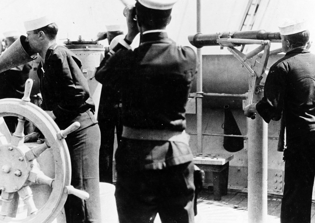 Scene on the ship's fire control bridge, 18 May 1918. Note man at speaking tube at left, officer with binoculars in the center, and telescope at right. (Naval History and Heritage Command Photograph NH 54660)