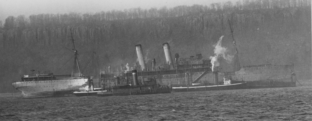 Listing slightly to port and attended by fireboats and tugs, DeKalb lies in the Hudson River near Spuyten Duyvil Creek, 16 December 1919, after she had been damaged by a fire that had broken out while the ship was lying ready to be converted to a...