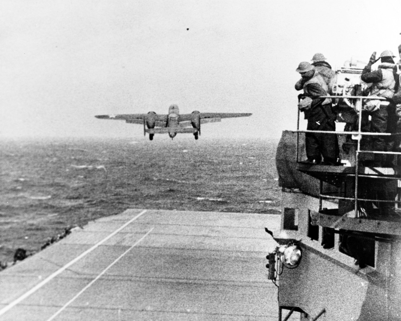 A B-25B Mitchell of the Army’s 17th Bombardment Group takes off from Hornet to bomb the Japanese home islands, 18 April 1942. Note the men wearing the old British style M1917A1 helmets as they watch from the signal lamp platform (right). (U.S. Na...