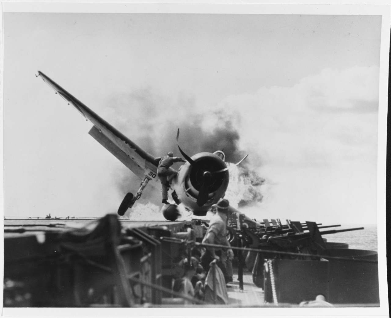An F6F-3 of VF-2, Ens. Byron M. Johnson, crashes into the ship’s port 20 millimeter gun gallery while landing and the Hellcat’s belly fuel tank erupts into flames, 10 November 1943. Lt. Walter L. Chewning Jr., USNR, the catapult officer, scramble...