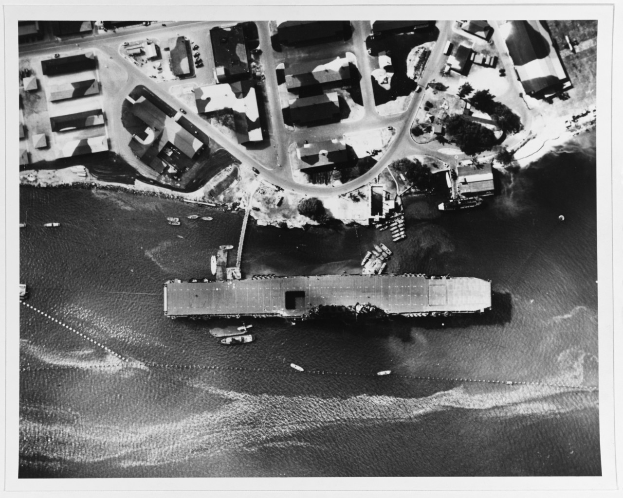 A plane captures this overhead view of the ship moored at Ford Island, 12 July 1942. Note the Wildcat on the barge alongside aft, the antitorpedo nets, and the camouflaged buildings on the island. (Naval History and Heritage Command Photograph NH...