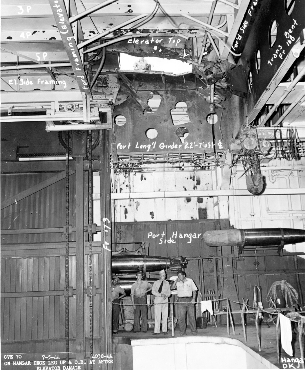 Fanshaw Bay at Pearl Harbor Navy Yard, 5 July 1944, showing a hole in the elevator (top) from a Japanese bomb. Note the size when compared to the man seen on the flight deck, and the size of the men on the hangar deck involved in inspecting the d...