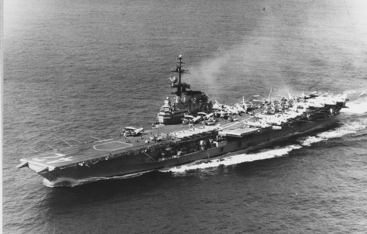 An aerial photograph of the ship captures her slicing cleanly through a calm sea, most likely during her deployment to the Vietnam War, 1966–1967. The Phantom IIs parked on the flight deck sport AB tail codes, identifying them as serving in CVW-1...