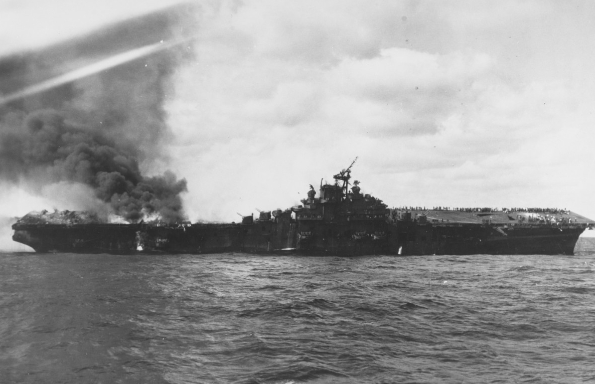 Franklin on fire and listing following the attack on 19 March 1945. Note what appears to be the towing wire from Pittsburgh (CA-72) attached to the outboard end of the carrier’s starboard anchor chain. Photographed from Santa Fe. (U.S. Navy Photo...