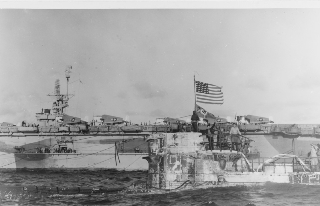 The U.S. flag flies over the German naval ensign from U-505’s periscope, which the boarders use as a jackstaff, 6 June 1944. This picture shows the submarine two days after her capture, and American sailors run hoses over the side to drain the wa...