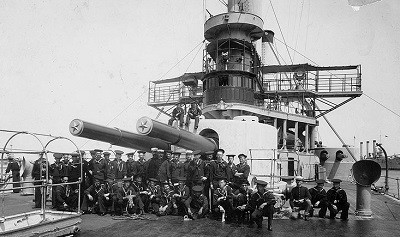 Sailors and marines of the ship’s company pose in front of her forward 12-inch gun turret, circa August 1898. Note that Iowa has been partially repainted from her wartime gray. Brooklyn (Armored Cruiser No. 3) lies in the distance (R). (Naval History & Heritage Command Photograph NH 00087, original photograph by William H. Rau)