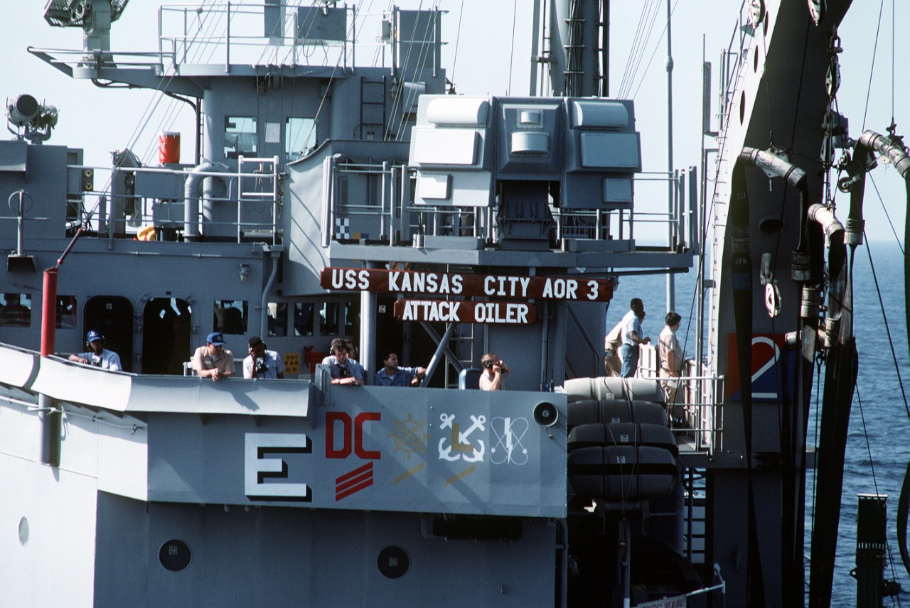 Officers and crewmen stand on Kansas City’s bridge wing during an underway replenishment, April 1991. Note the award symbols painted on the hull, including the large white E signifying the Battle E Award, which Kansas City won for the year 1990. ...