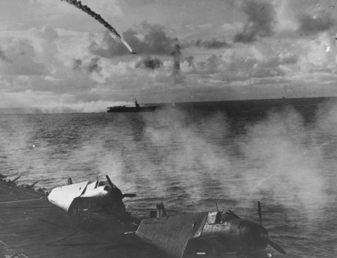 A Japanese twin-engine bomber crashes in flames as it attacks the escort carriers, 18 June 1944. Photographed from Kitkun Bay, with two of her Avengers, wings folded, parked on the flight deck, and guns blaze against the assailing enemy aircraft....
