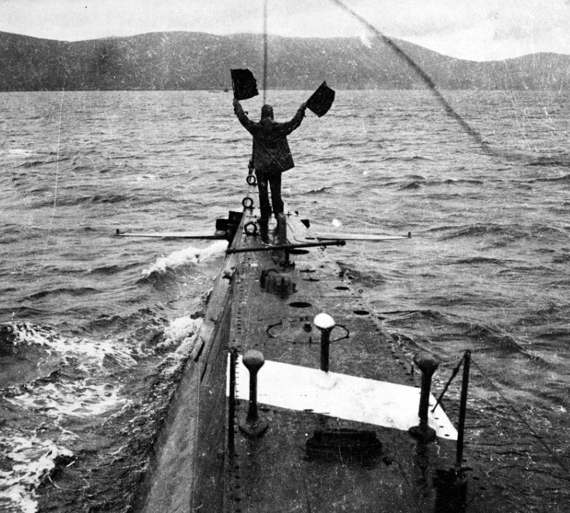 AL-3 crewman sends a message to another submarine (visible in the center distance) using semaphore flags, during operations off Berehaven in 1918. Note SC Tube type hydrophone and white stripe (probably a recognition marking) on AL-3's foredeck. ...