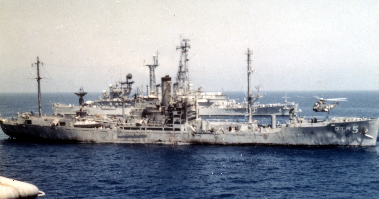 Liberty, her appearance reflecting the ferocity of the attack by Israeli planes and motor torpedo boats the previous afternoon, lists to starboard while Little Rock passes in the background and a helicopter lifts off to transfer wounded to Americ...