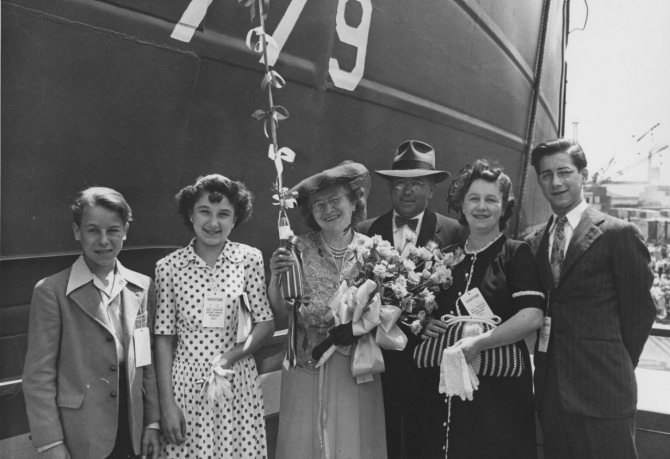 Mrs. Andrew Vavrek, LST-779’s sponsor (center) holding the traditional bottle and bouquet, beams for the camera with others of the launching party before christening the ship that looms behind her, 1 July 1944. (U.S. Navy Bureau of Ships Photograph 19-N-67841, National Archives and Records Administration, Still Pictures Branch, College Park, Md.)