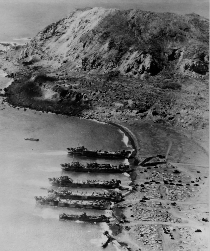 “... And the unloading of general cargo is proceeding”. Five tank landing ships and one medium landing ship nose into the dark sands of Iwo Jima on 24 February 1945. The view shows Beach Green 1, with Mount Suribachi in the background. The small wrecked ship in the foreground is Japanese. The landing ships include (from bottom to center): LSM-264, LST-724, LST-760, LST-788, LST-808 (with a LCT embarked), and LST-779 (carrying a pontoon causeway). (U.S. Navy Photograph NH 65314, Naval History and Heritage Command)