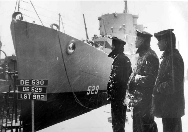 Three Mason crewmembers admire their ship while she lies moored at the Boston Navy Yard, Mass., on 20 March 1944. The placards indicate that Mason’s sister ship John M. Bermingham (DE-530) and the tank landing ship LST-982 are fitting-out nearby....