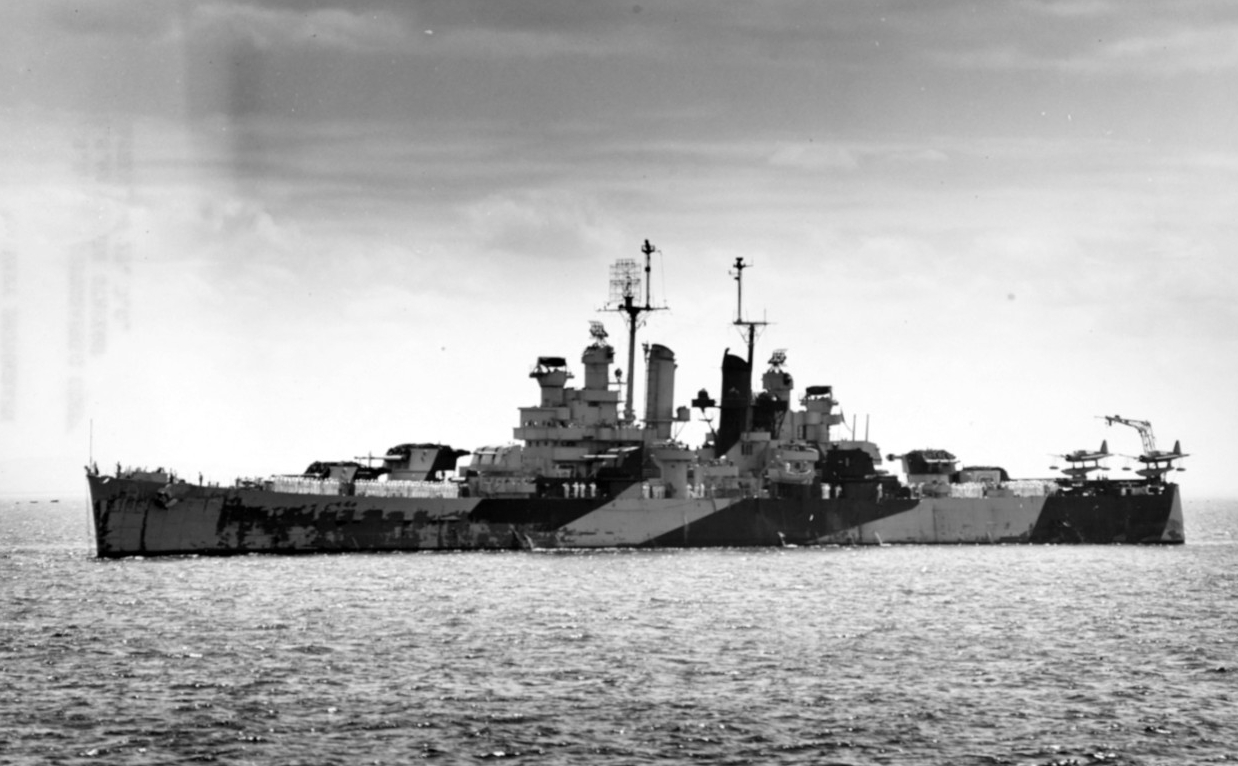 Miami at Trinidad during her shakedown, 19 February 1944, photographed from Quincy. Note her crew at quarters on deck, the very weathered paint of her Measure 32, Design 1D camouflage on her hull, and a pair of Vought OS2U-3 Kingfishers of Cruise...