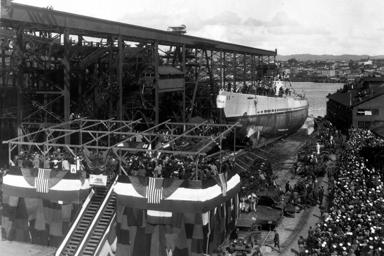 V-6 slides down the ways at Mare Island while onlookers crowd almost every conceivable space – ranging from rooftops to building ways, even to the maintop of the heavy cruiser Chicago (CA-30) under construction in the next building ways at left –...