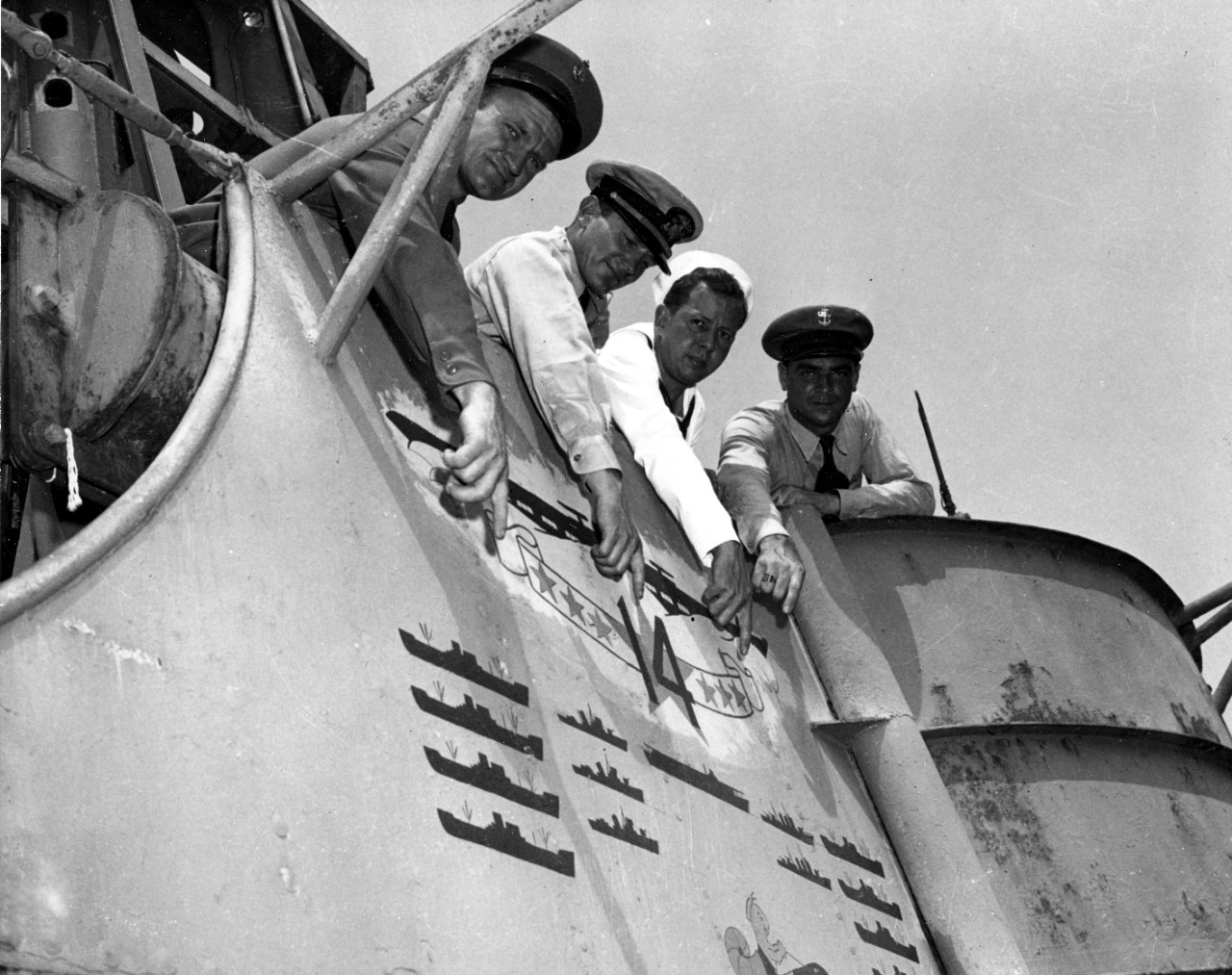 Unidentified members of Nautilus’s crew point proudly to the “scoreboard” painted in the boat’s conning tower, 26 January 1946. Note cartoon insignia almost out of view at the bottom of the image, of a smiling aquatic creature wearing a “dixie cu...