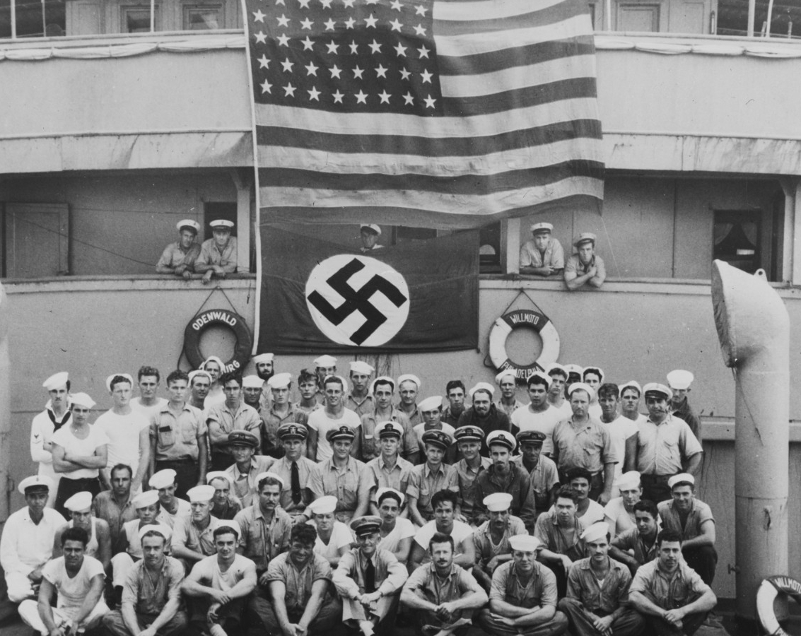 Omaha’s boarding party, Lt. George K. Carmichael (in the center with white cap cover), poses beneath the Stars and Stripes displayed over the German flag on board the captured blockade runner Odenwald, 18 November 1941. Note life rings ODENWALD H...