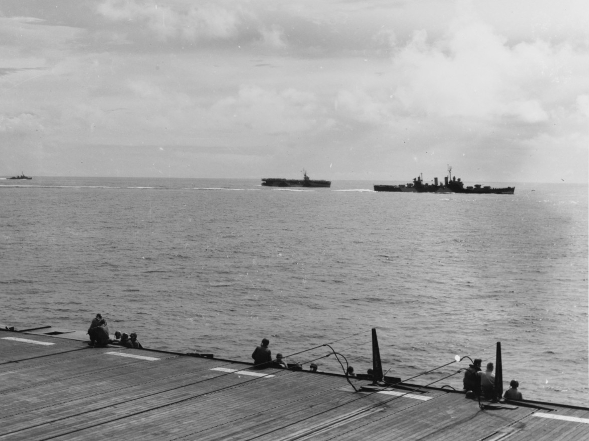 A carrier crewman snaps this shot of a starboard beam view of Phoenix (right) as she screens escort carriers off Leyte, 30 October 1944. Note the flight deck sailors gazing at the ships arrayed, and the barriers rigged in the foreground. (U.S. Na...