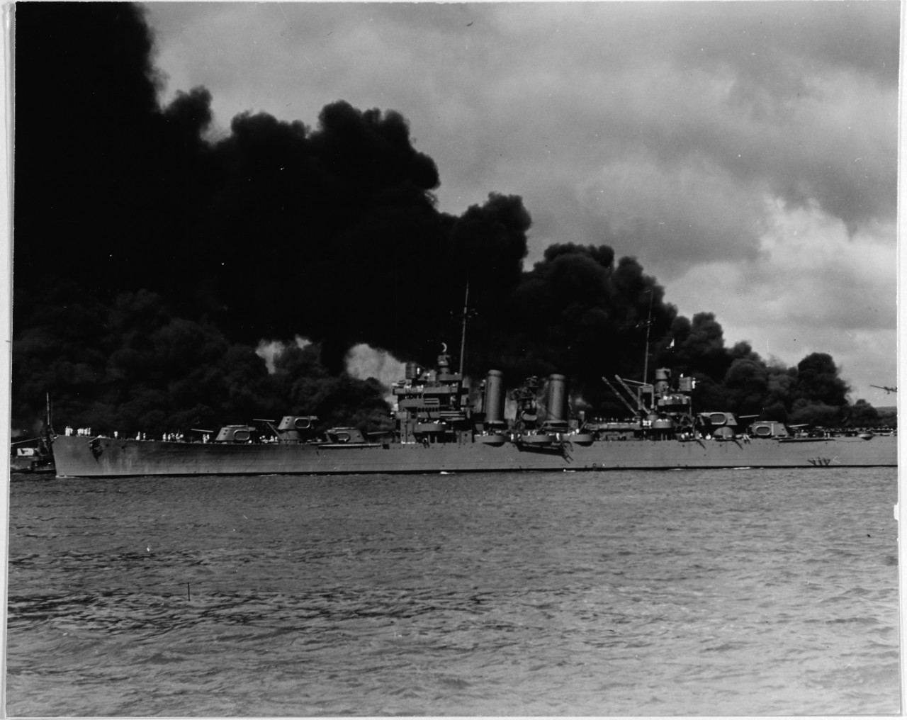 Phoenix steams past burning West Virginia (BB-48) (left) and Arizona (BB-39) (right) along the south side of Ford Island as she sorties during the Japanese attack on Pearl Harbor, T.H., 7 December 1941. (Naval History and Heritage Command Photogr...