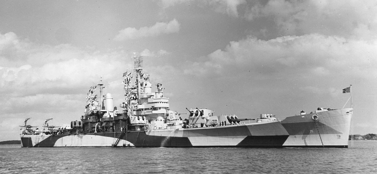 Quincy wraps-up her availability at Boston Navy Yard, 14 April 1944. The ship breaks out an array of signal flags and appears in a camouflage scheme, bristling with radar and radio antennae, and embarks a pair of Kingfishers spotted on the catapu...