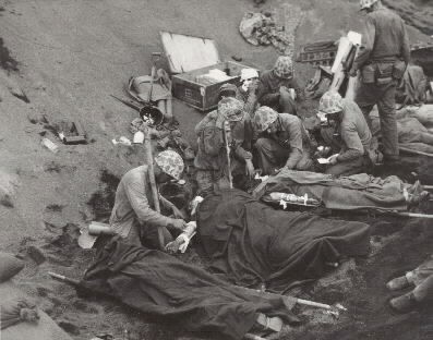 Iwo Jima Invasion, February 1945: Navy doctors and corpsmen administer to wounded marines at an Iwo Jima first aid station, 20 February 1945. Navy Chaplain, LtJG John H. Galbreath (right center) is kneeling beside a man who has severe flash burns...