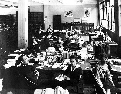 Navy Department, Washington, D.C. Military and civilian personnel in an office in the Main Navy or Munitions Buildings, circa 1919. Most of those present are Yeomen (F). A Medical Corps Lieutenant is seated in the front left center, dictating to a Yeoman (F). Note white shield around the mouthpiece of the telephone in the center of the photo; also wire document baskets on desks and Liberty Loan posters on the wall