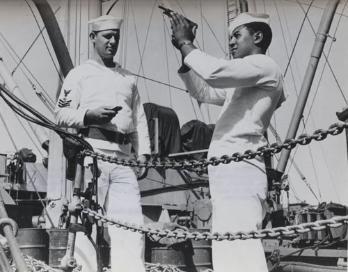"Relieving the watch at the quarterdeck somewhere in the Pacific, Coxswain William Green observes safety precautions in checking his pistol while Albert S. Herbert, Quartermaster First Class, stands by with a clip of ammunition and holster belt, ready to complete the formalities."